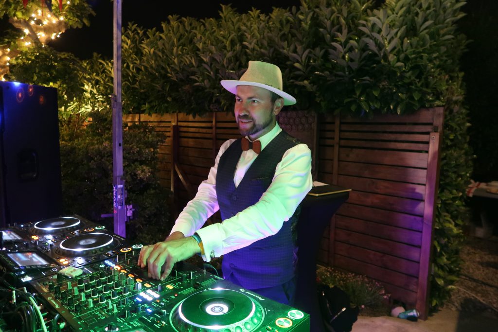 Mężczyzna (DJ) w kapeluszu, białej koszuli, kamizelce i muszce, obsługujący podświetloną konsoletę didżejską. Zdjęcie wykonane w nocy, w tle widać ogrodzenie i rośliny.