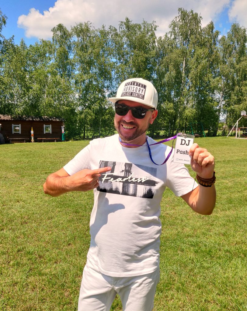 Mężczyzna (DJ Pash), ubrany w białą koszulkę, czapkę z daszkiem i okulary przeciwsłoneczne, uśmiecha się i wskazuje palcem na identyfikator z napisem "DJ Pash!" zawieszony na szyi. Znajduje się na zewnątrz, na tle zielonej trawy i drzew.
