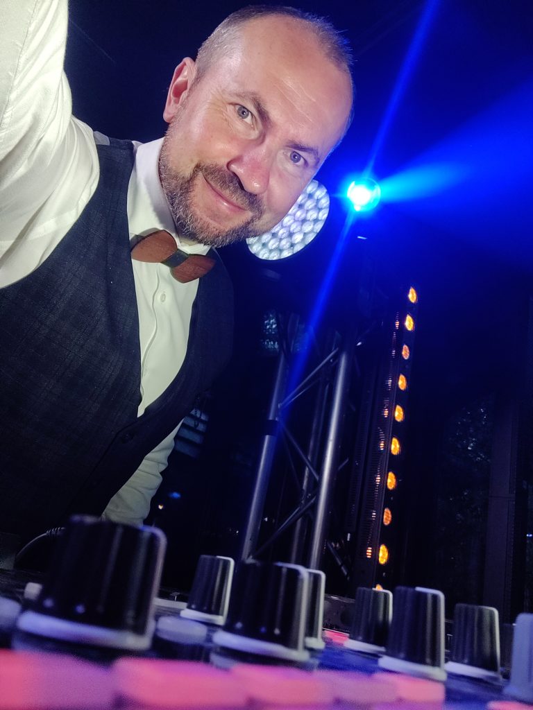 Uśmiechnięty mężczyzna, DJ, ubrany w białą koszulę i kamizelkę z drewnianą muchą, ujęty w zbliżeniu od dołu, zza konsolety. Za nim widać reflektory sceniczne emitujące niebieskie światło.